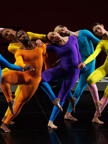 Seven dancers dressed in single-colored, tight-fitting jumpsuits, all in the same position, standing on one leg with the other leg bent back.