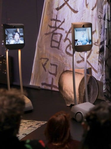 3 robots écrans qui montrent 3 visages de hikokomori. Les robots sont face au public, vu de dos, asssis sur le sol.