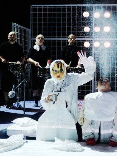 deux personnages en costumes blancs, l'un masqué et l'autre ressemblant à une marionnette, devant des musiciens