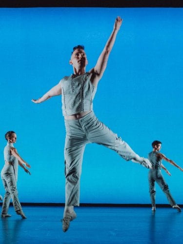 Four dancers in light-colored costumes in front of a vibrant blue background