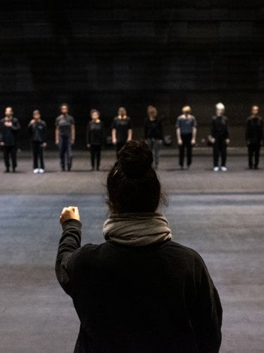 Nacera Belaza from behind, and facing her, 12 dancers in a line dressed in black