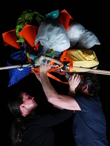Two people supporting a large, colorful, and varied load of objects