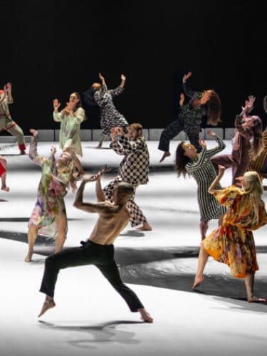 group of dancers in diverse, colorful costumes on a white and black stage, performing