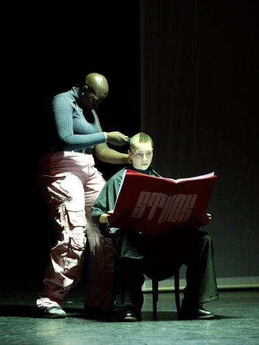 one dancer shaves the head of another who is seated and reading a large red book with "STUCK" written on it