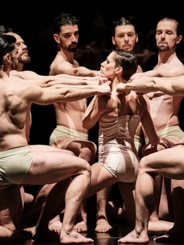 Shirtless dancers, one knee on the ground, forming a circle around a female dancer, their arms extended towards her