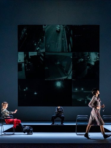 five dancers on stage, a row of black folding chairs, and a large screen displaying a grid of dark surveillance video feeds