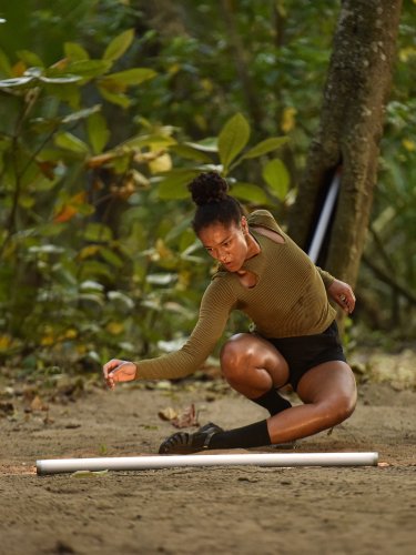 Dancer crouching on earthy ground in a lush forest, arm extended towards a white tube placed on the ground