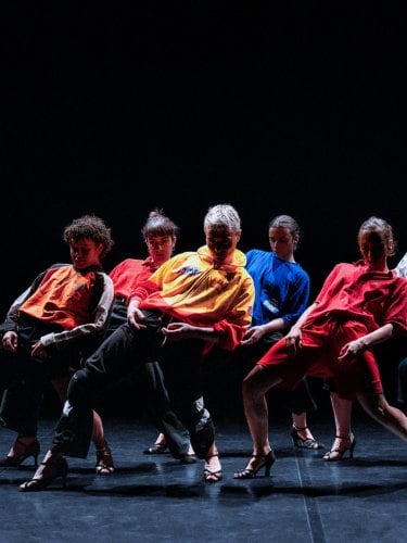 Group of dancers in colorful clothing and high heels striking dynamic poses on a dark stage