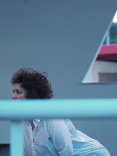 Curly-haired woman in white shirt looking right, with triangular window and greenery in background