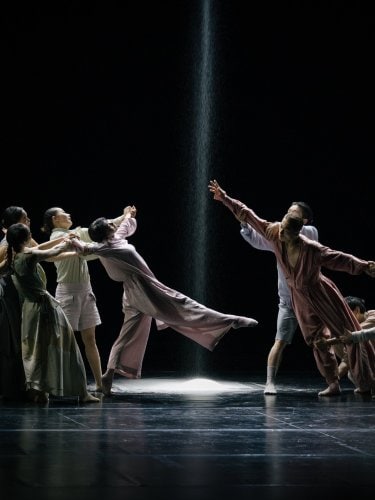 Dancers in flowing costumes on a dark stage, with a beam of light and falling sand in the center.