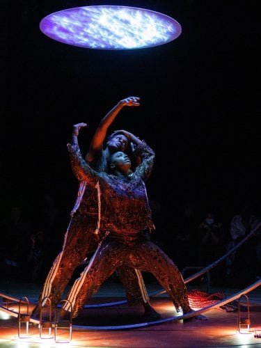 Deux danseurs regardent vers un plafond lumineux, leur corps embrasse un même mouvement dans une sphère métallique. 