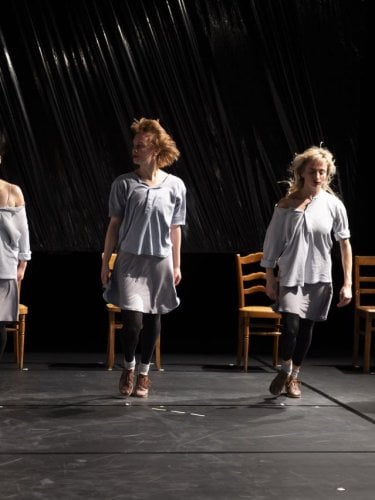 Quatre danseuses se tiennent debout alignées devant 4 chaises, avec un arrière fond noir. 