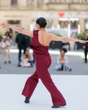 Dancer in a maroon outfit, seen from behind with outstretched arms, performing outdoors