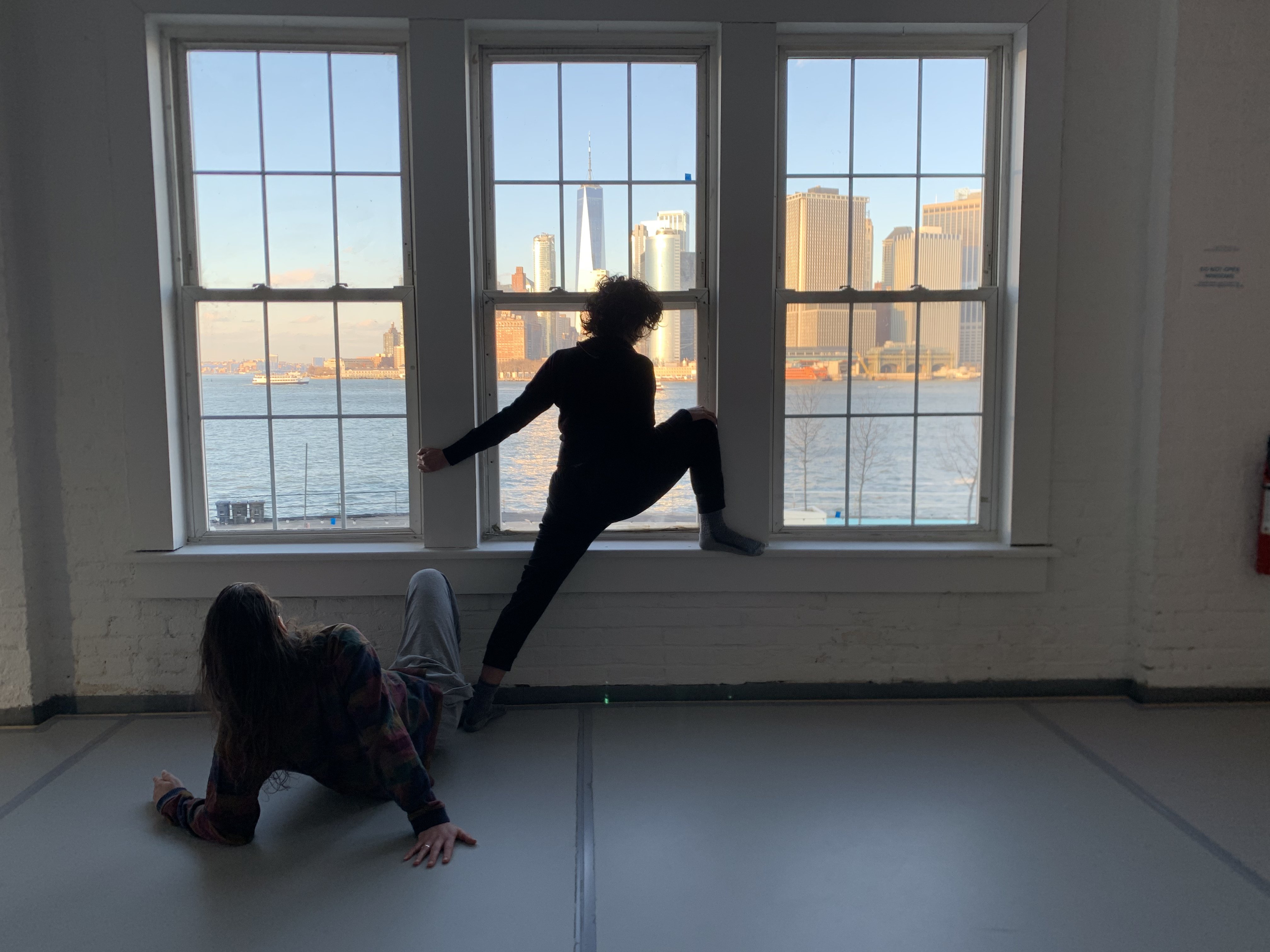 Two dancers stretching in front of a window with a view of  the New York landscape