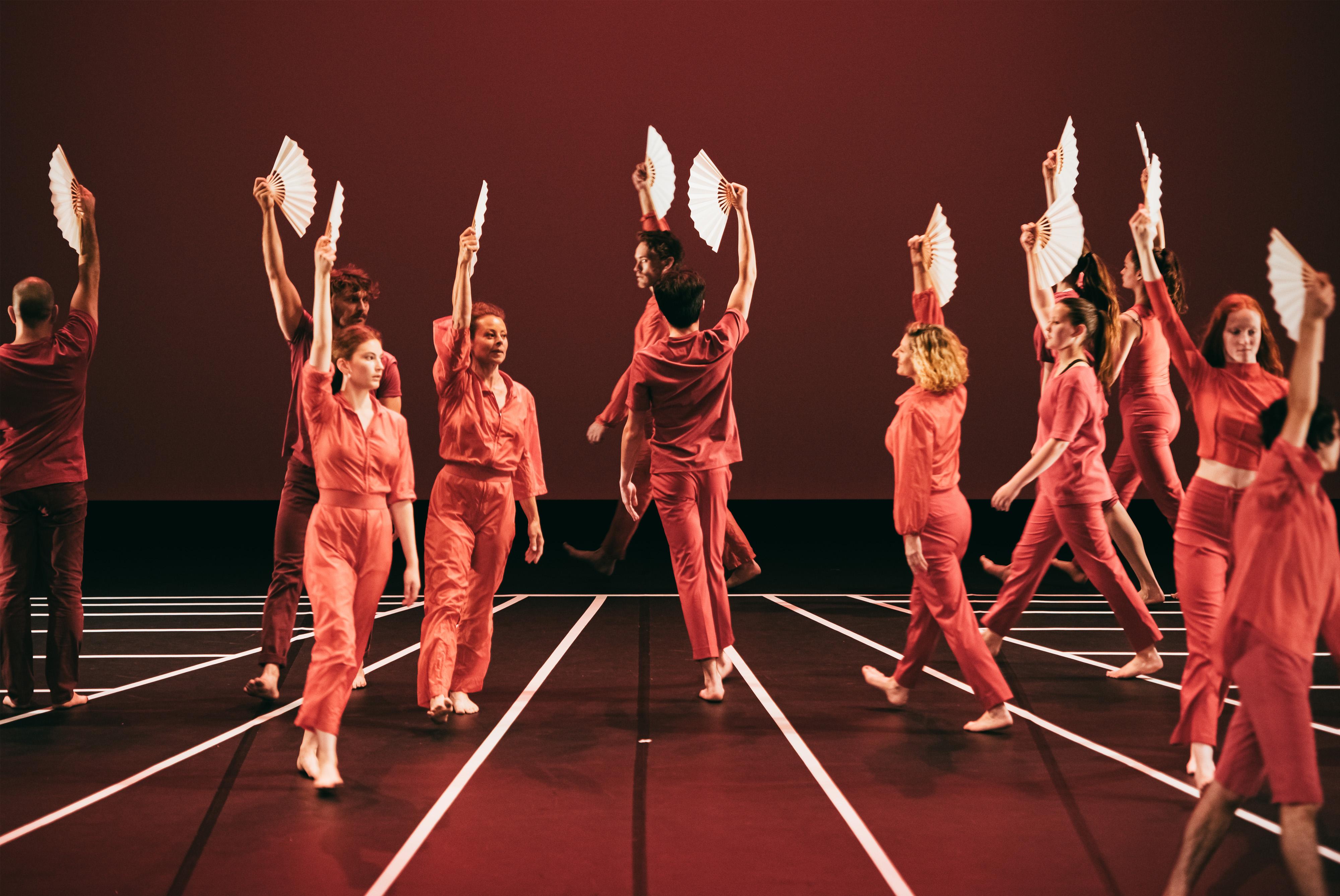 Dancers dressed in red, holding white fans above them, performing Andy De Groat's Fan Dance on the black stage covered with white stripes.