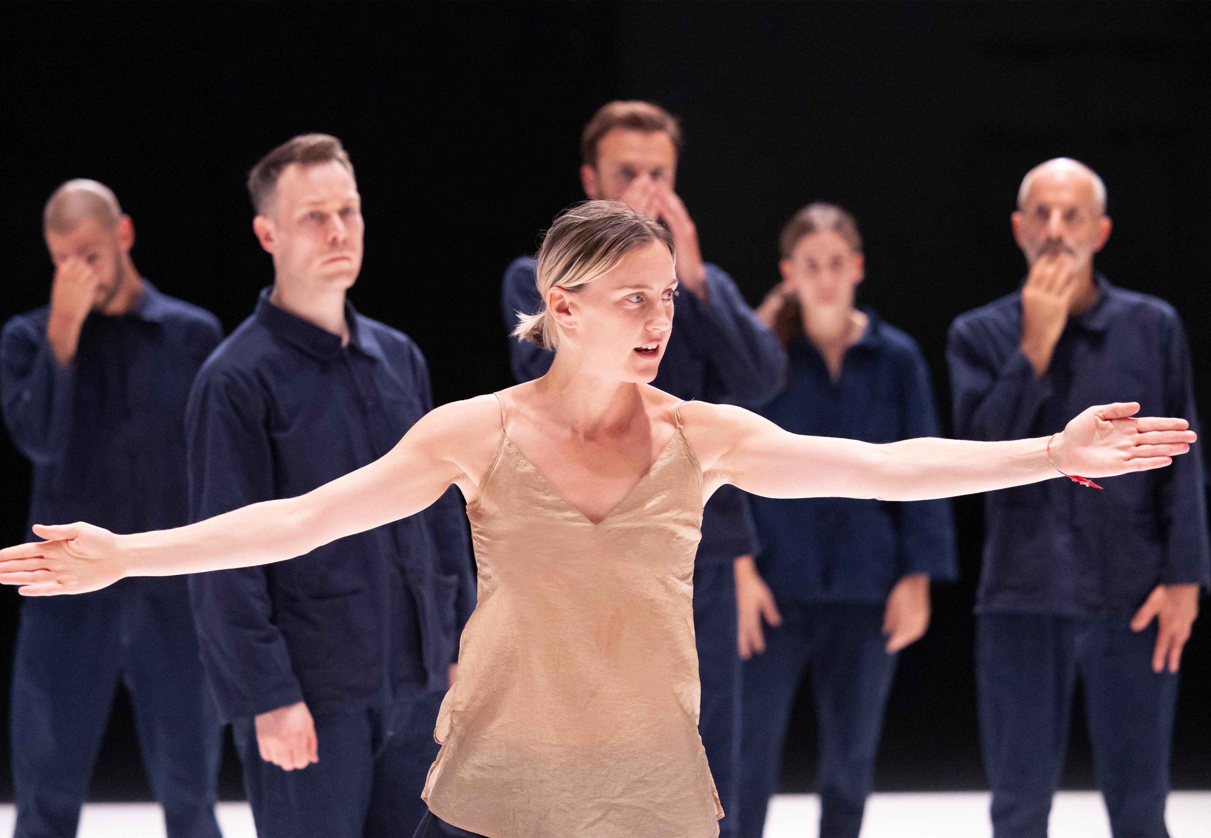 : Oona Doherty wearing a beige tank top, front, arms open with dancers dressed in overalls behind her.