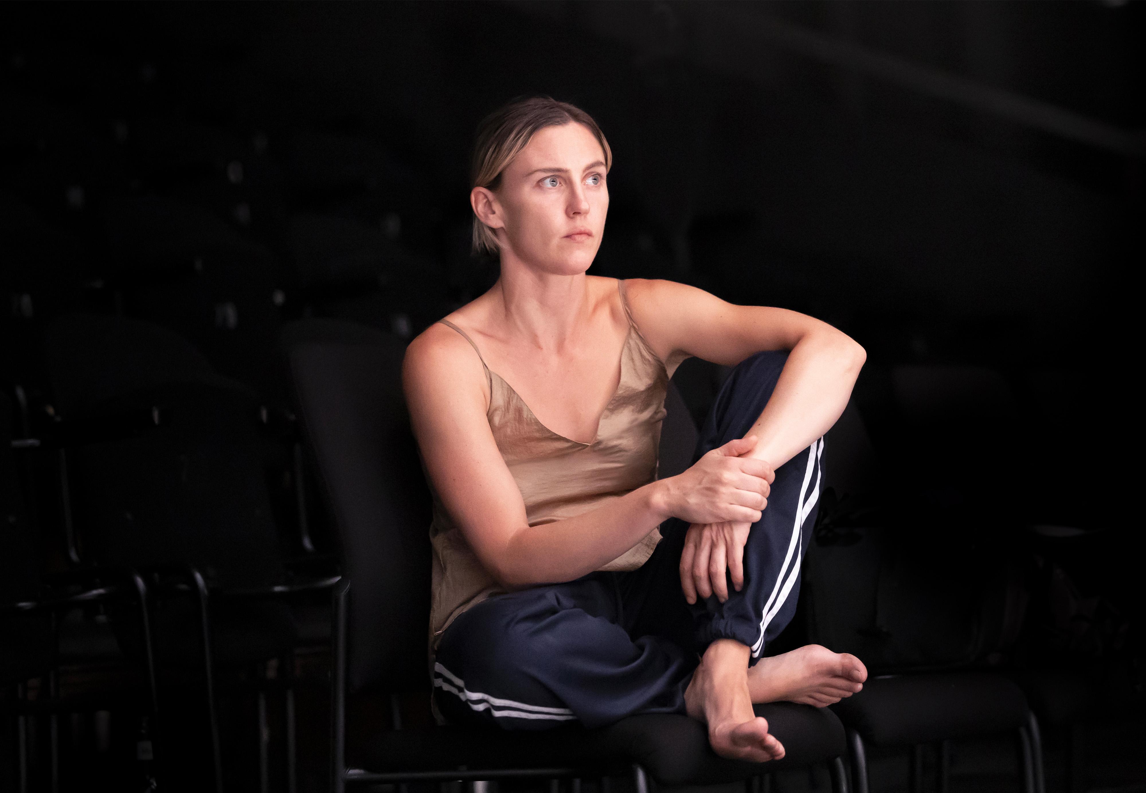 Portrait of Oona Doherty sitting cross-legged on a chair.