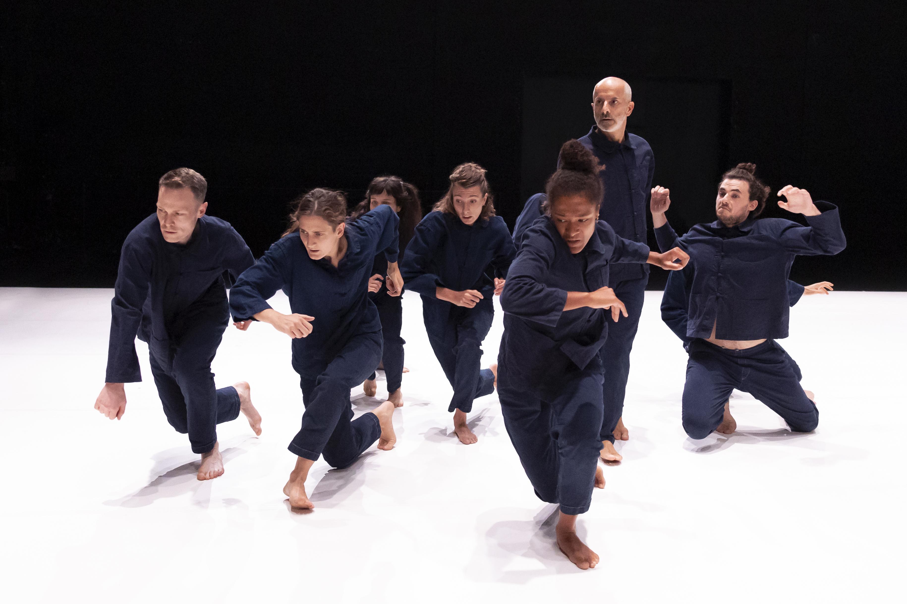 danseurs vêtus de bleus de travail, groupés et accroupis, les mains jointes sur les genoux