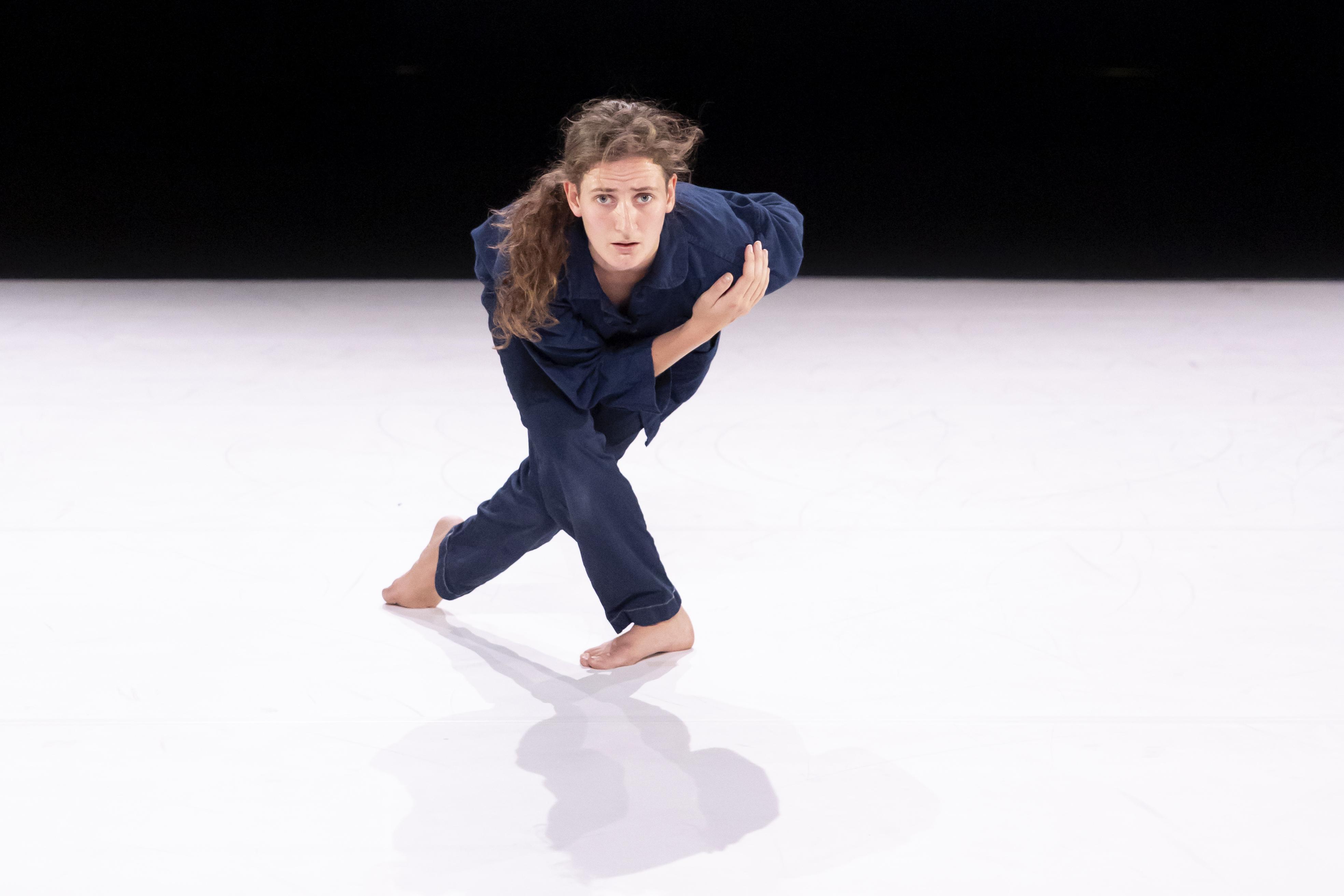 dancer dressed in overalls, facing forward, one leg crossed backwards and one arm on her chest 