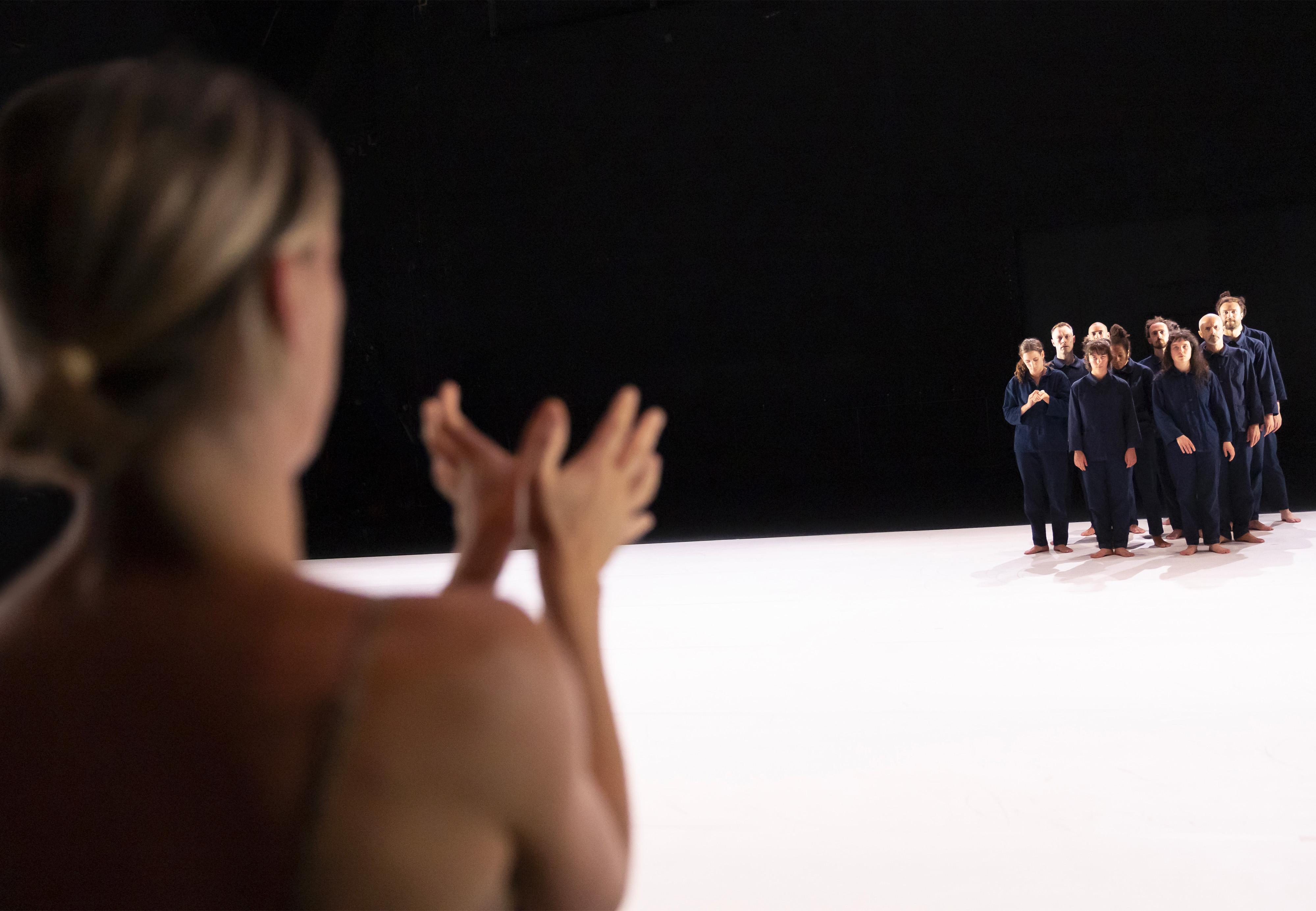 Oona Doherty, from behind, leading dancers dressed in overalls, grouped at the back of a stage covered with white linoleum.