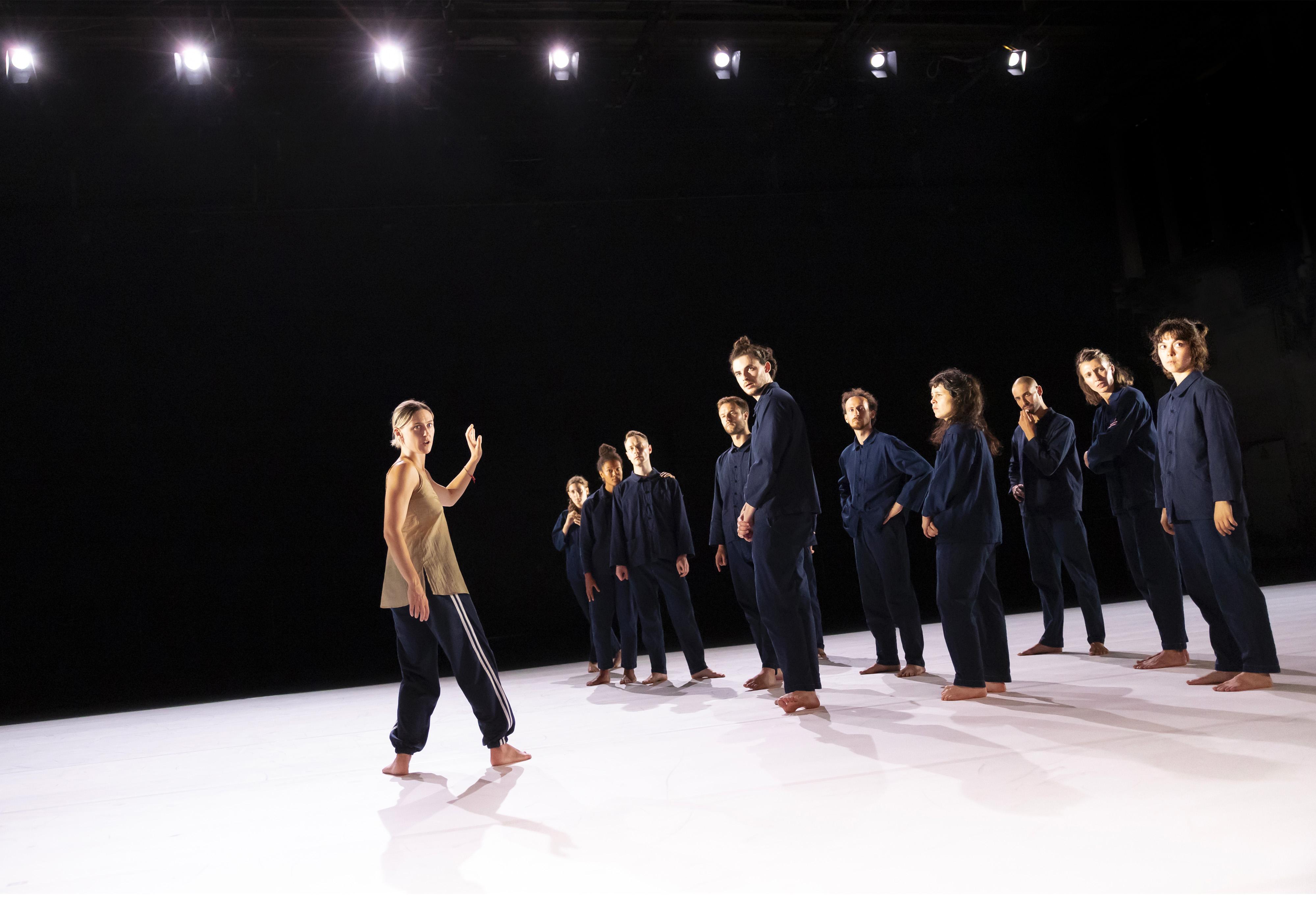 Oona Doherty, in profile, walking towards dancers dressed in overalls, grouped at the back of a stage covered with white linoleum.