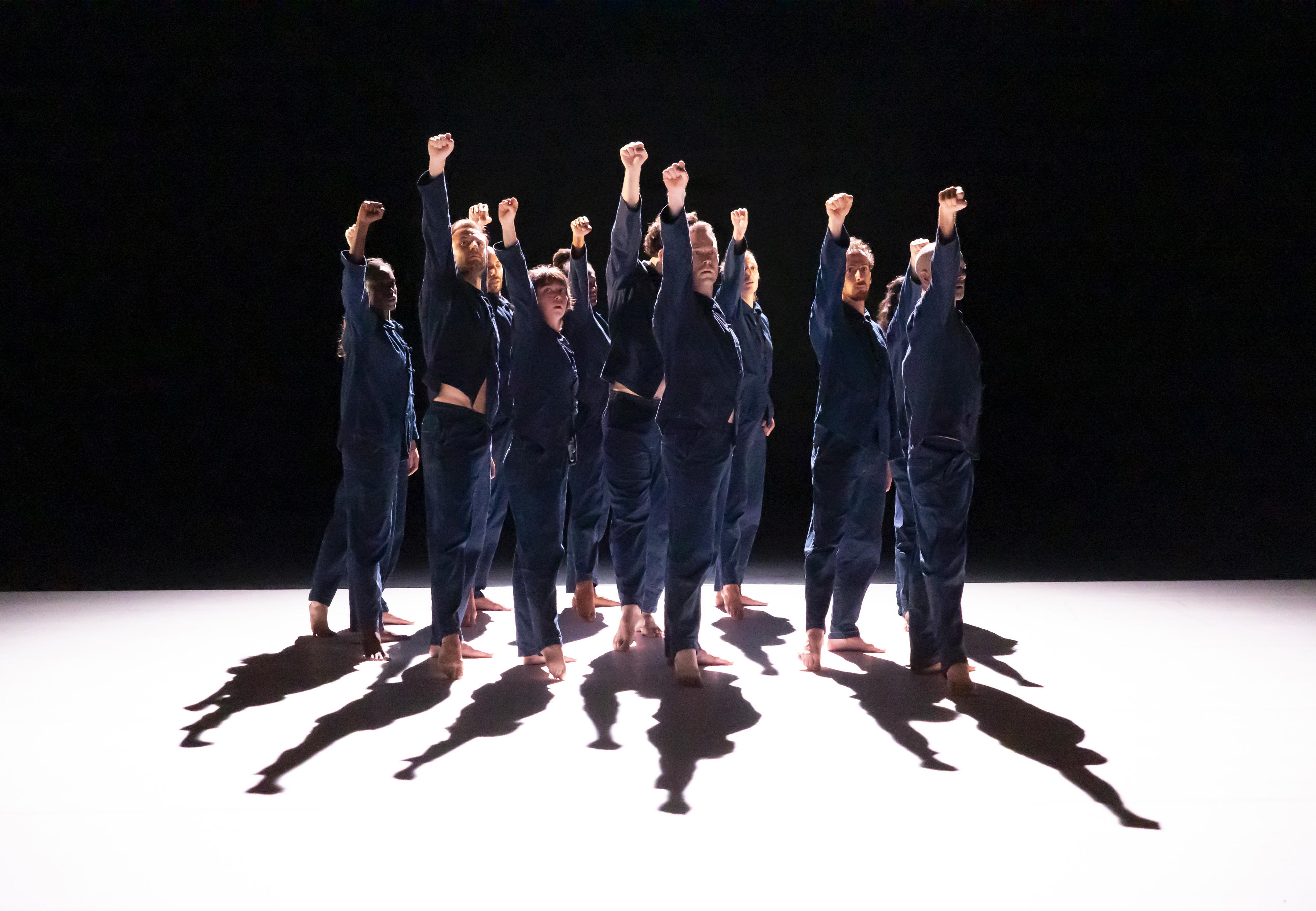 dancers dressed in overalls, grouped at the back of a stage covered with white linoleum, fists raised upwards.