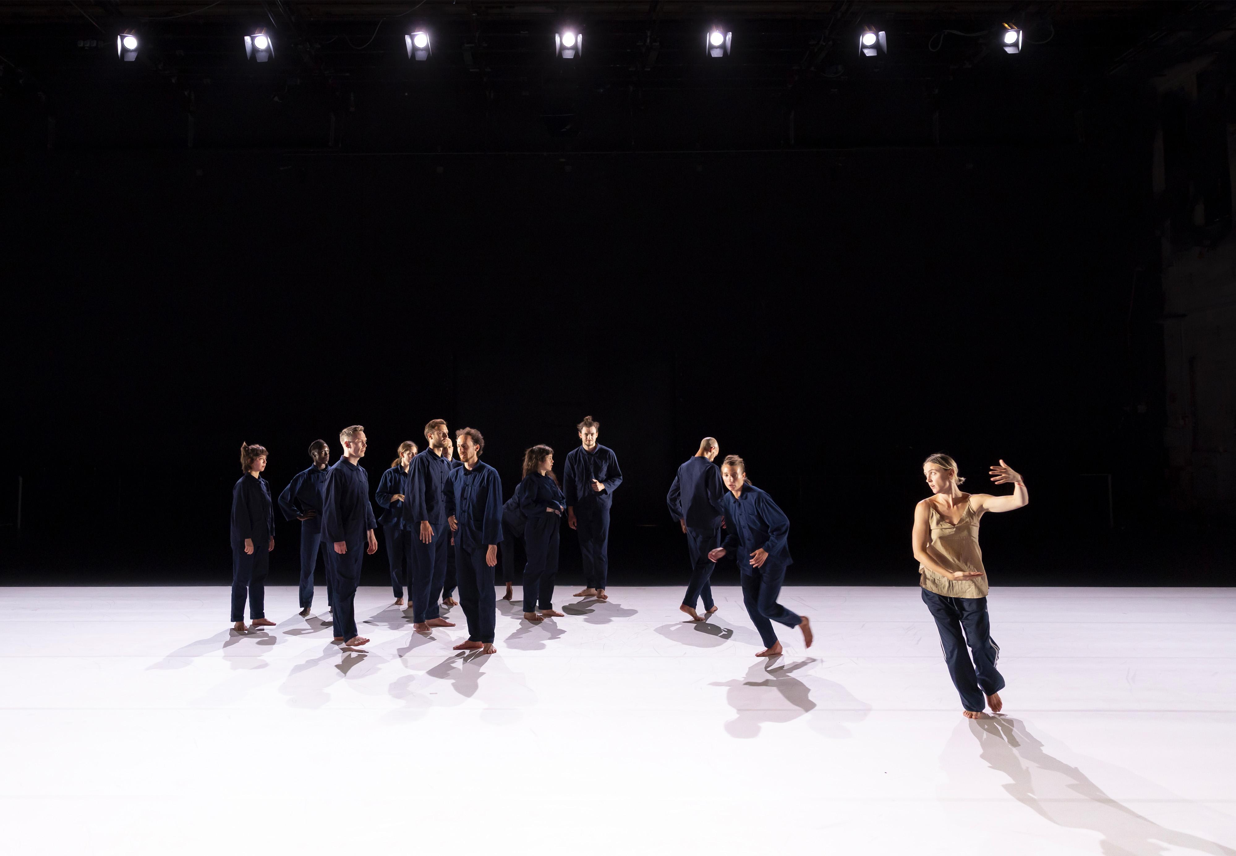 Des danseurs vêtus de bleus de travail sur une scène recouverte de lino blanc et Oona Doherty qui danse sur le côté avec un débardeur beige.