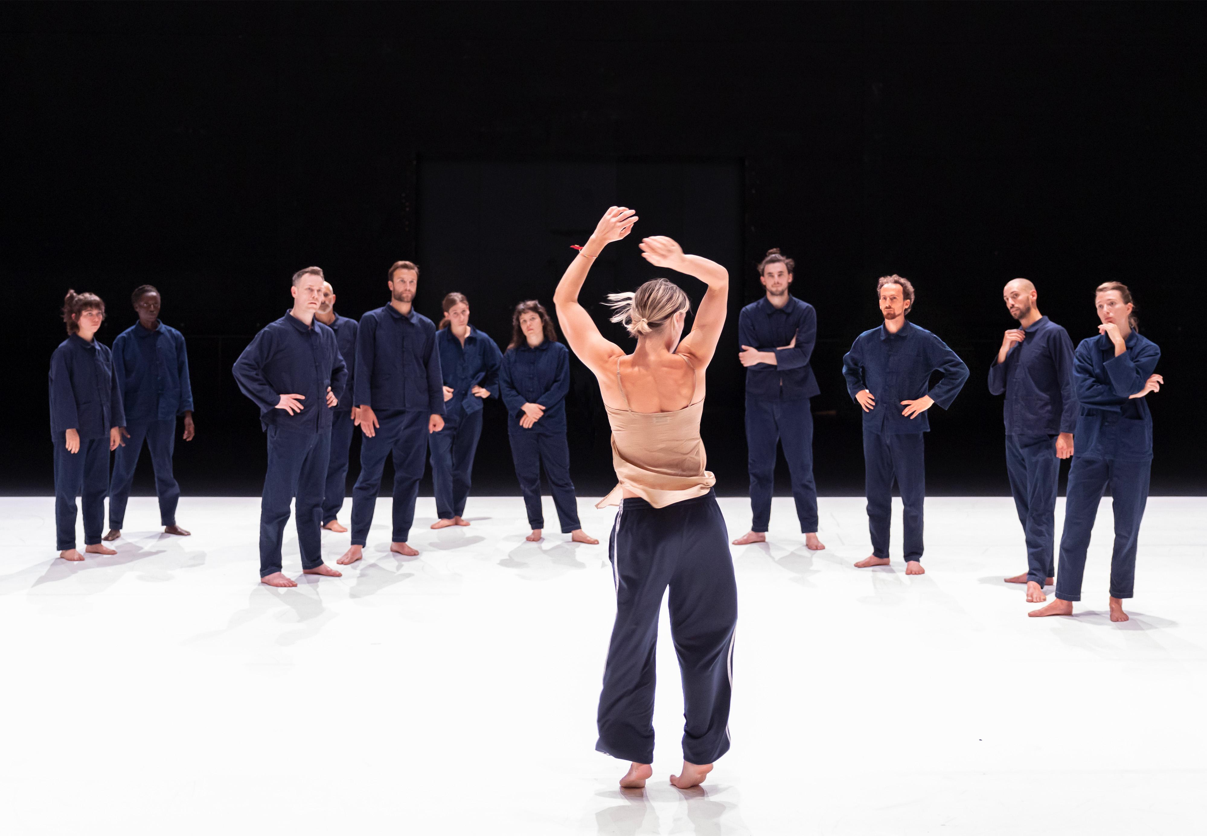 Des danseurs vêtus de bleus de travail sur une scène recouverte de lino blanc et Oona Doherty de dos qui danse face aux danseurs avec un débardeur beige