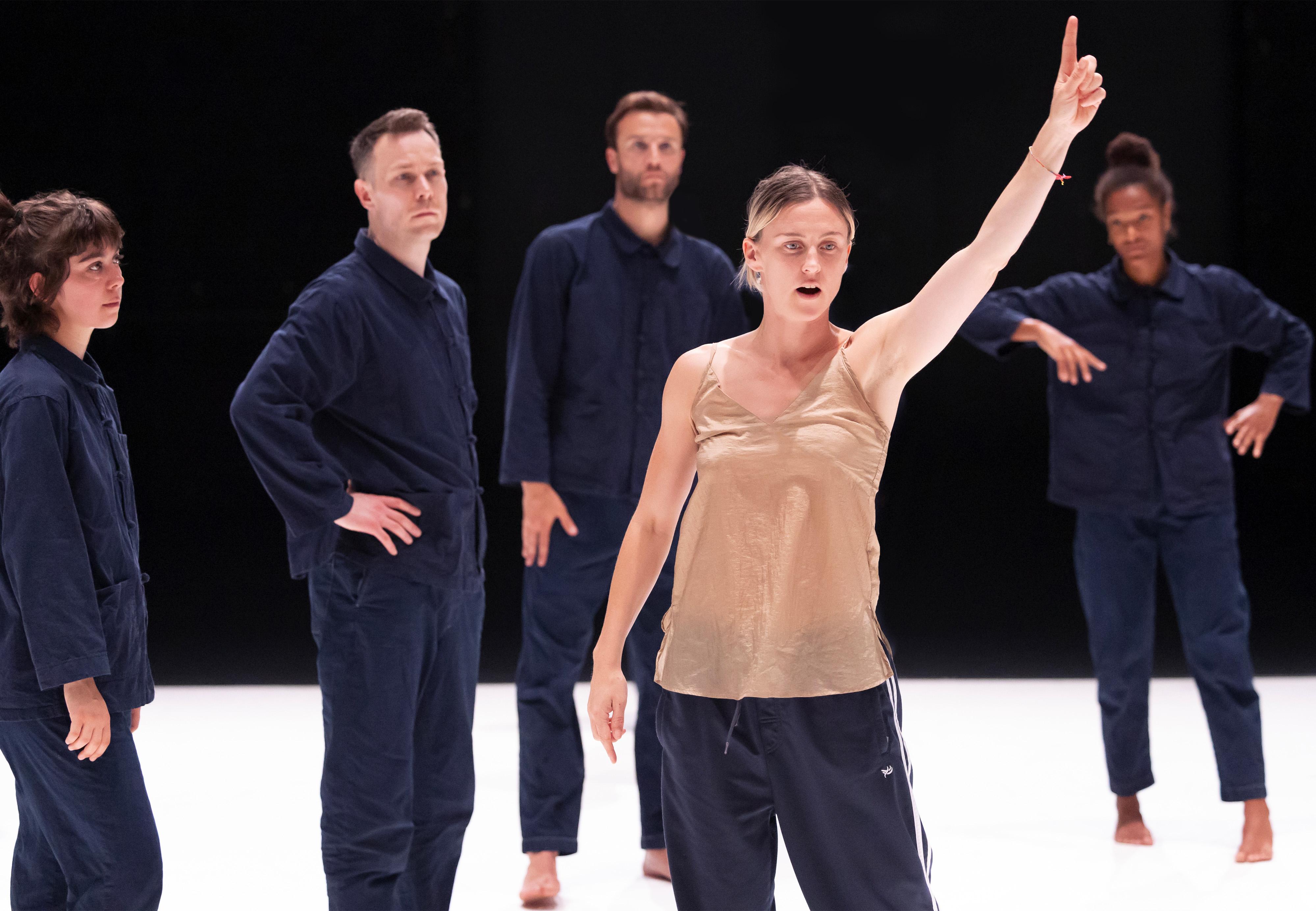 Oona Doherty portant un débardeur beige, de face, levant le bras vers le haut avec des danseurs vêtus de bleus de travail derrière elle.