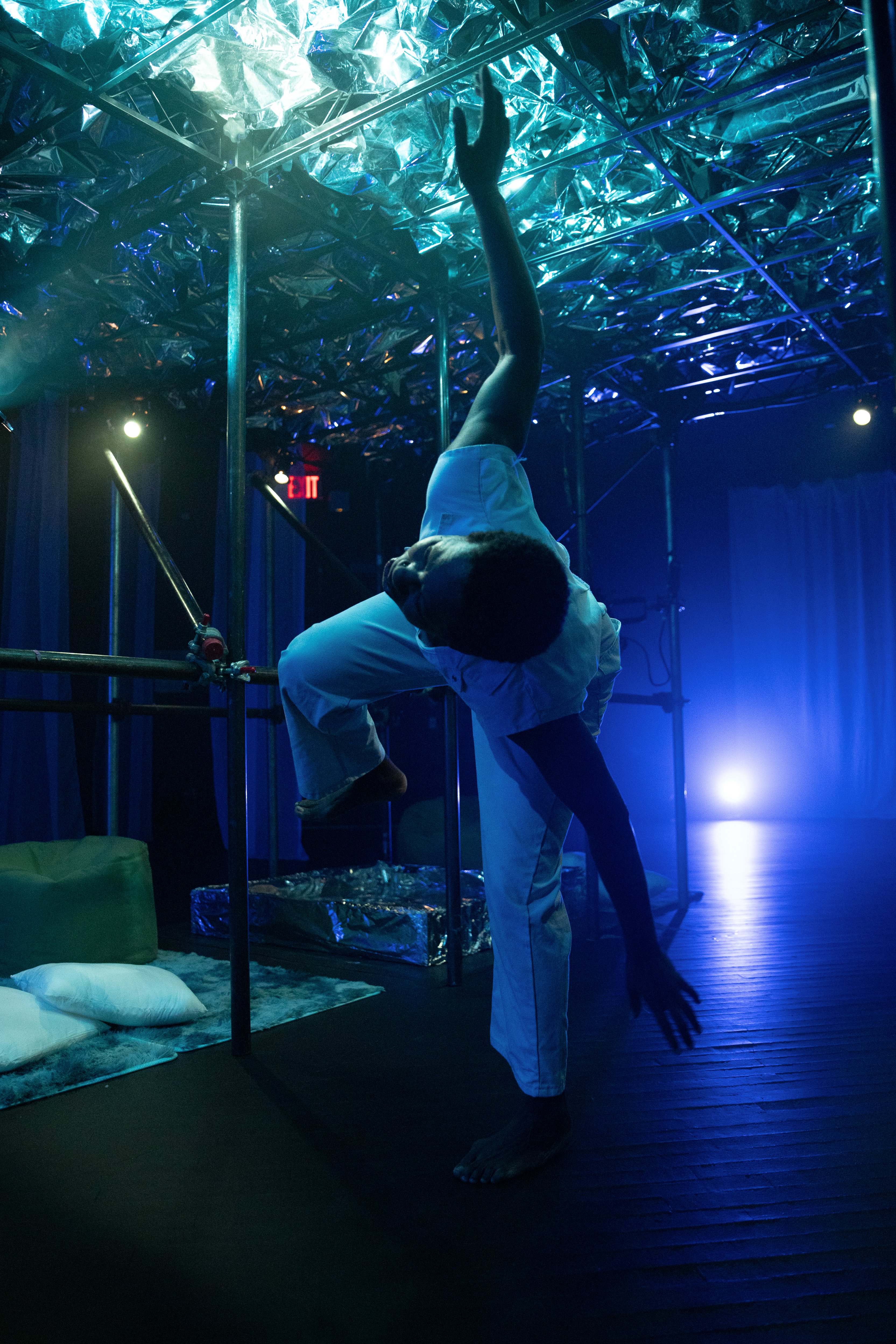 Mayfield Brooks is bent to one side with their arms outstretched, balancing on one leg. They are washed in the glow of a blue spotlight. 