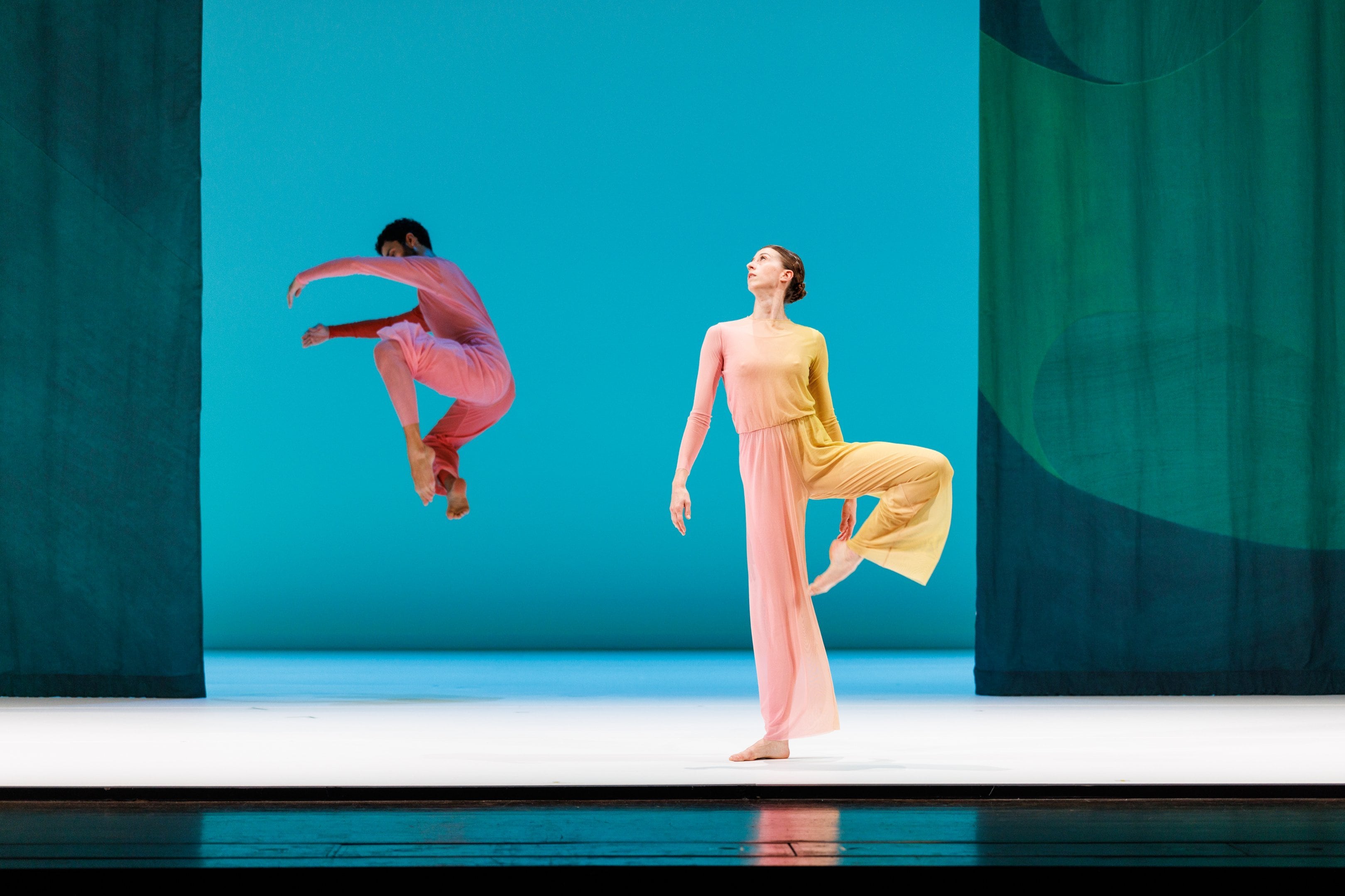 "A dancer in a pink and yellow costume and a dancer in a pink-orange costume jumping