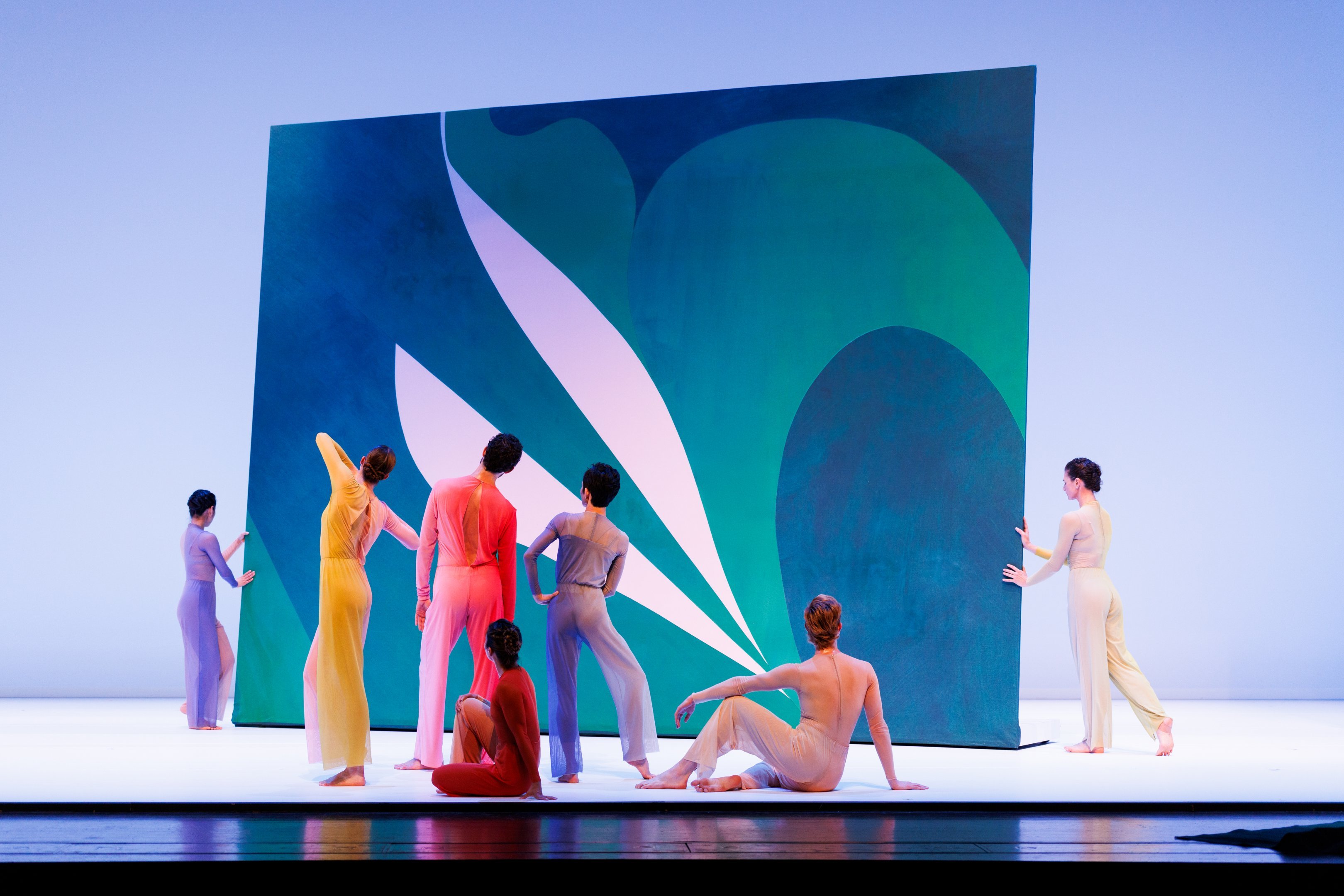 A group of dancers standing, one on the ground, all facing towards the back, facing a giant painting