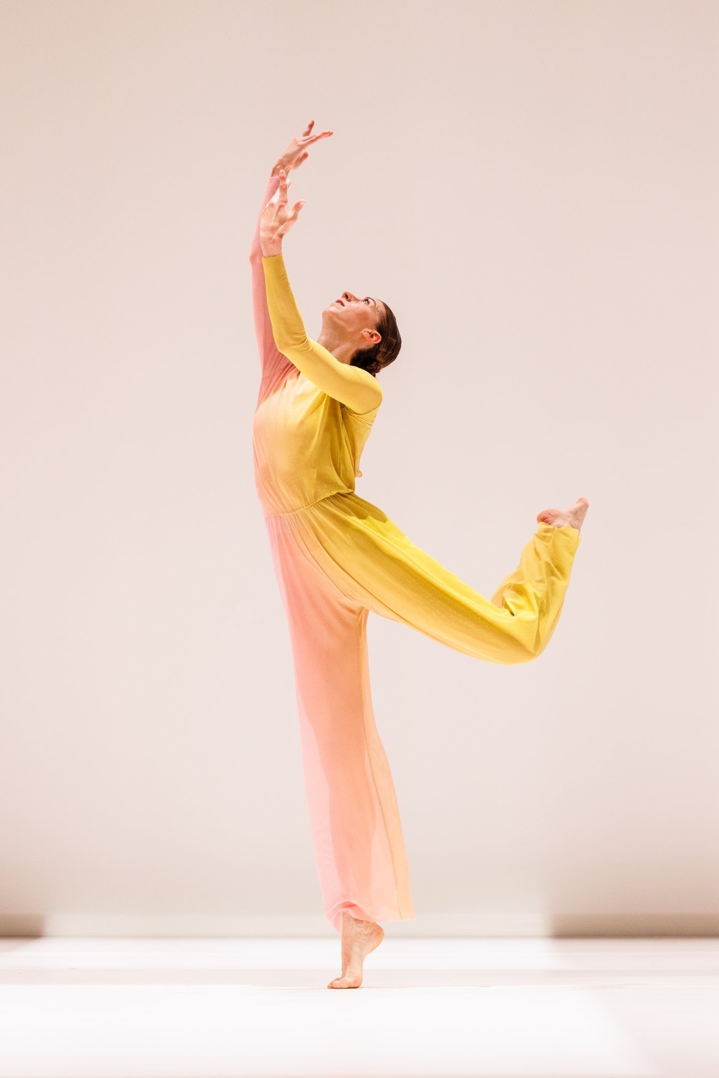 A dancer in a yellow and pink costume, right leg in attitude, and arms raised upwards