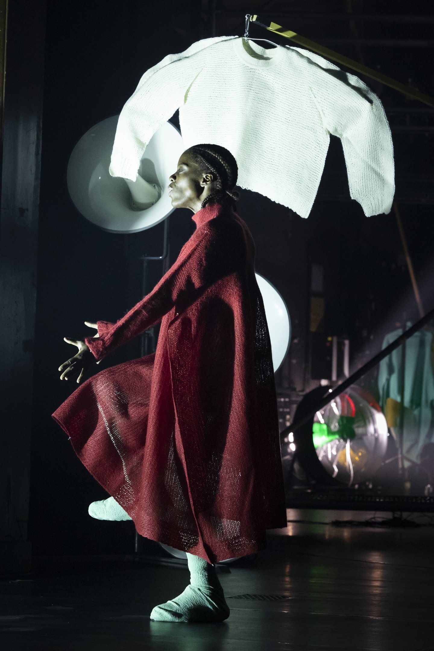 Dancer wearing a long, flowing red coat, a white knitted XXL sweater in the background
