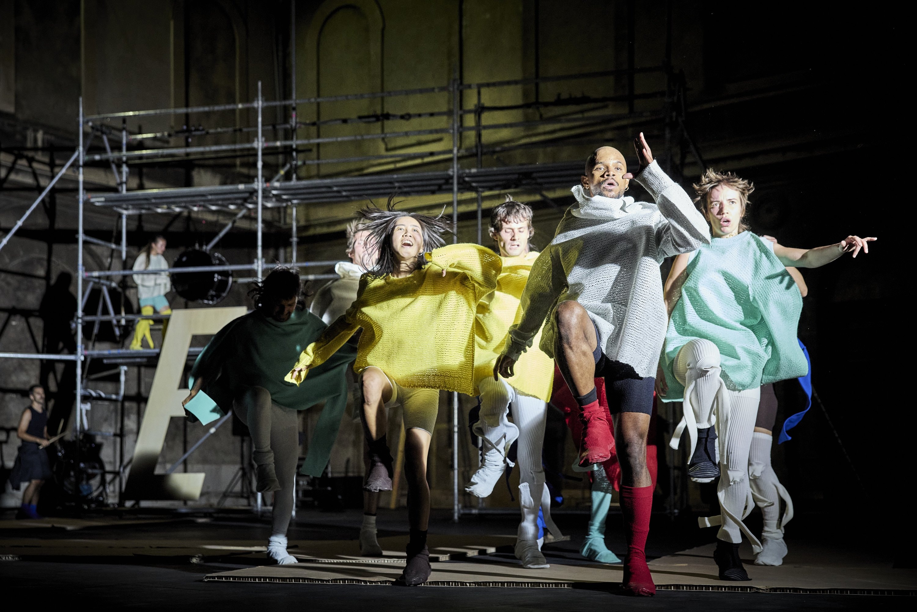 groupe of dancers in loose-fitting, brightly colored knitted tops and various bottoms are performing on a stage, captured in dynamic motion with expressive faces