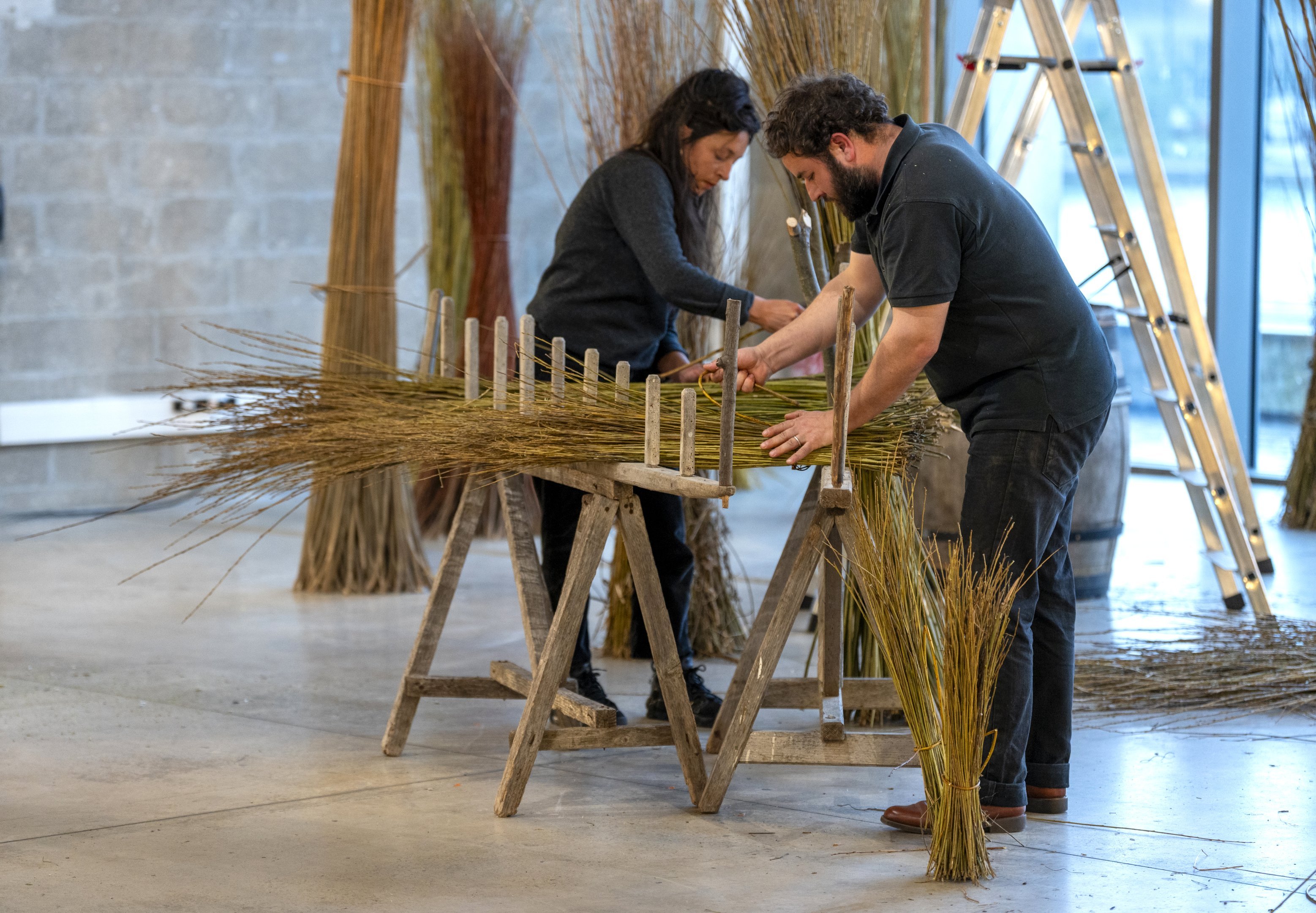 A man and a woman working with dried grasses on a workbench