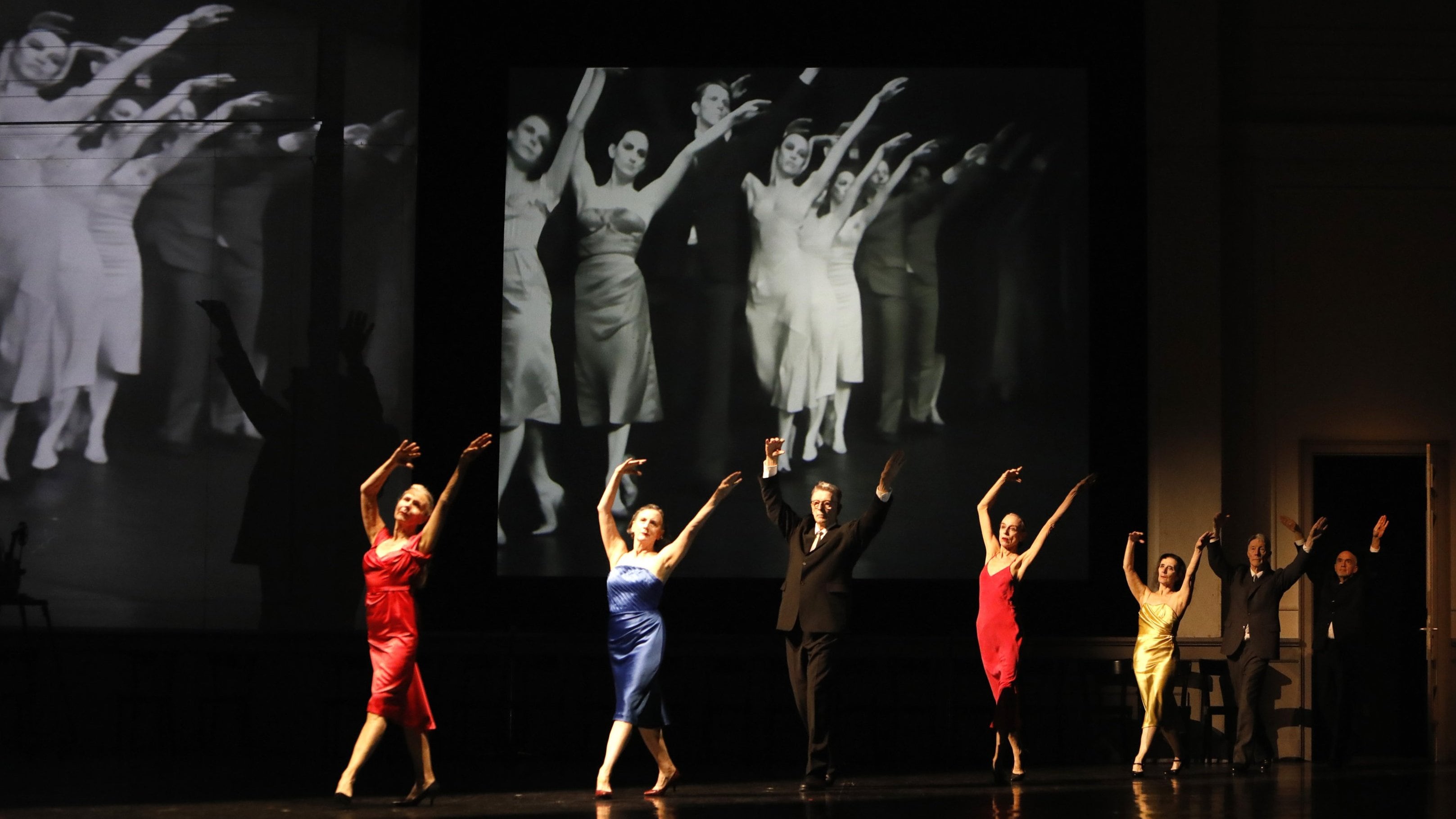 Quatre femmes et un homme en position statique avec un écran au fond qui montrent des danseurs dans la même position. 