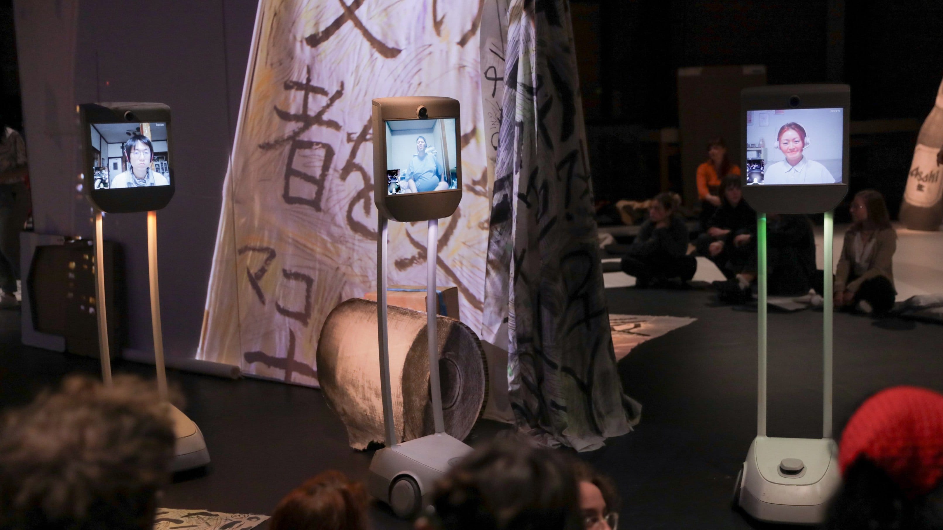 3 robots écrans qui montrent 3 visages de hikokomori. Les robots sont face au public, vu de dos, asssis sur le sol.