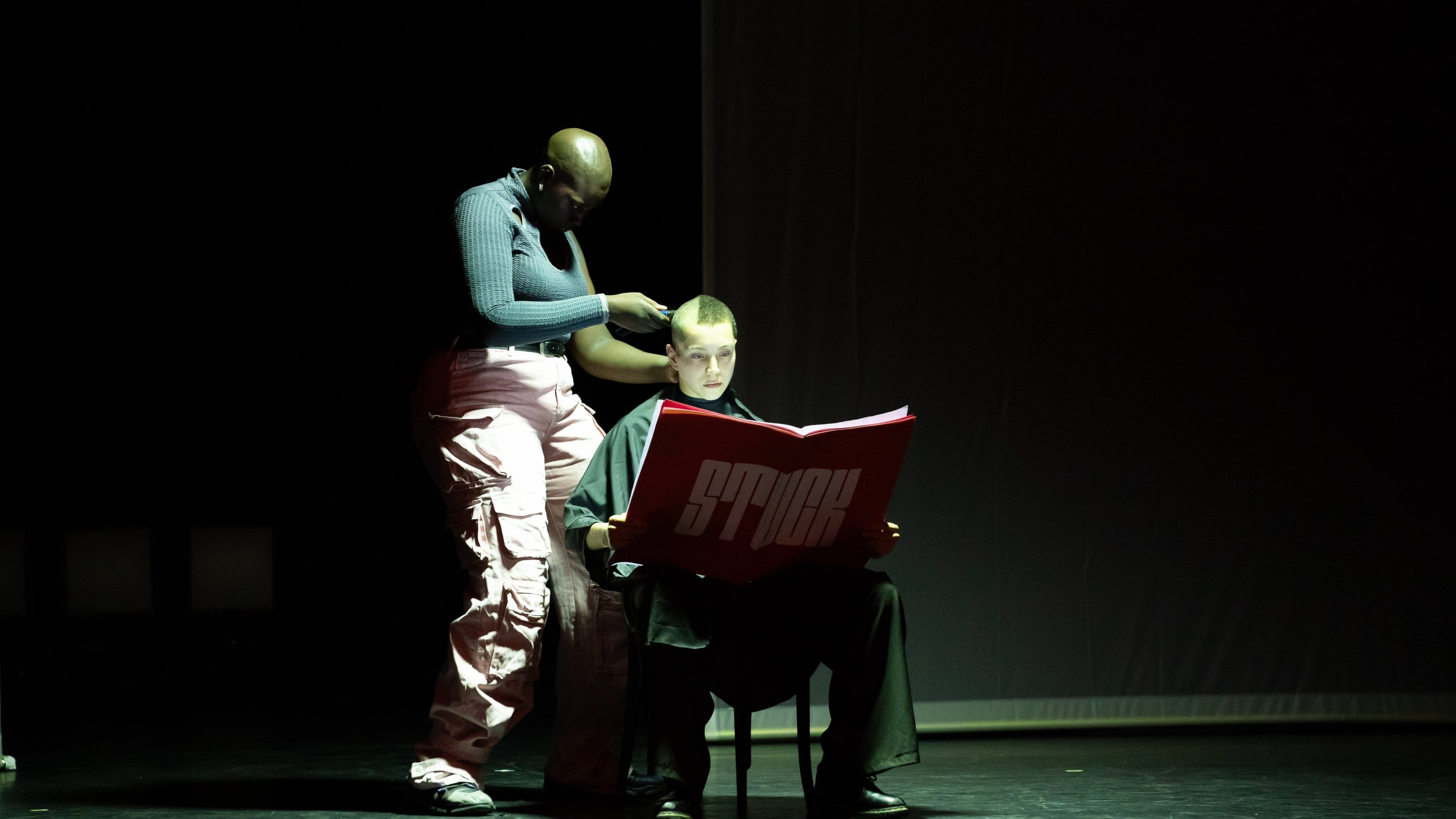 one dancer shaves the head of another who is seated and reading a large red book with "STUCK" written on it