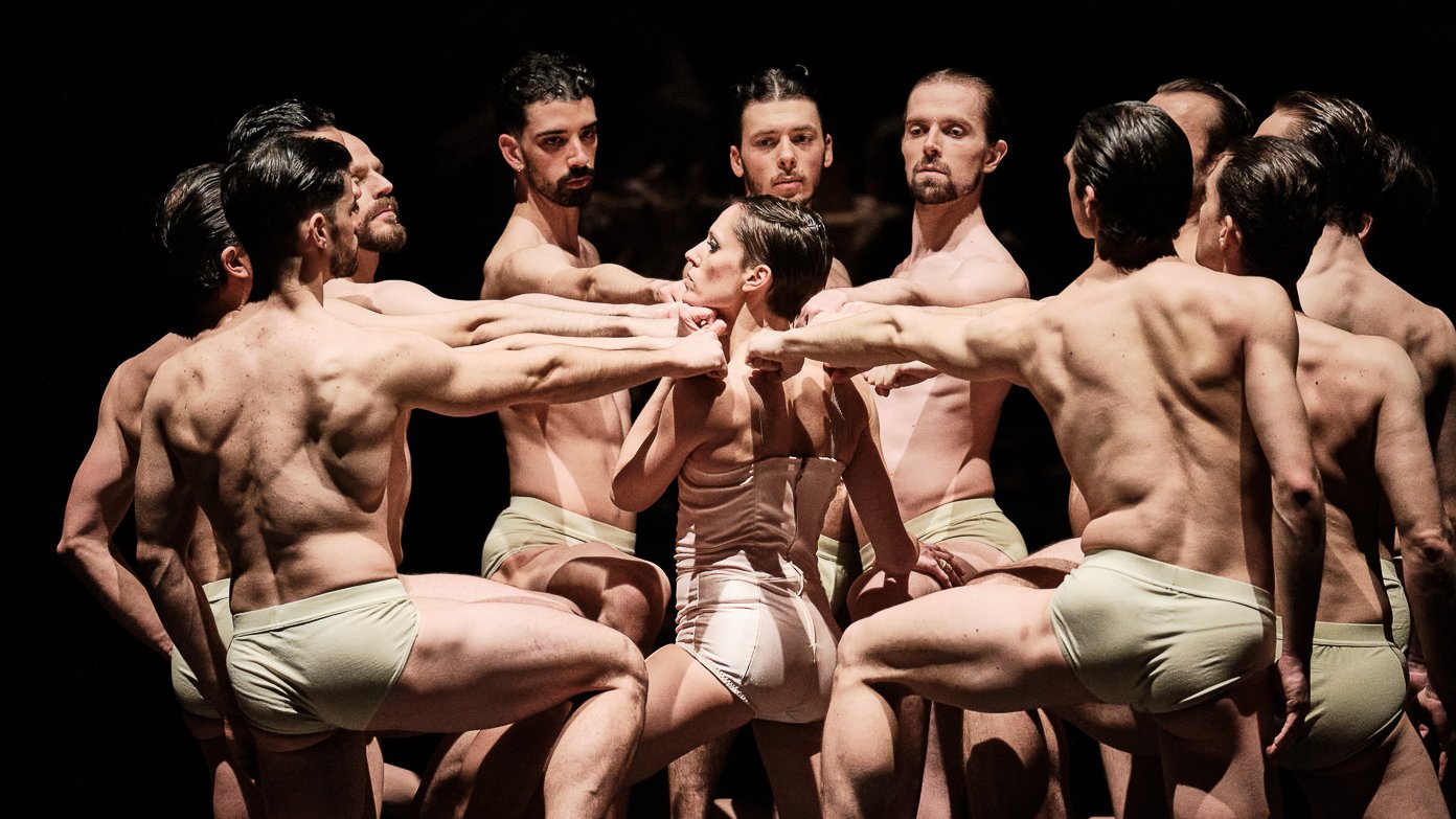Shirtless dancers, one knee on the ground, forming a circle around a female dancer, their arms extended towards her