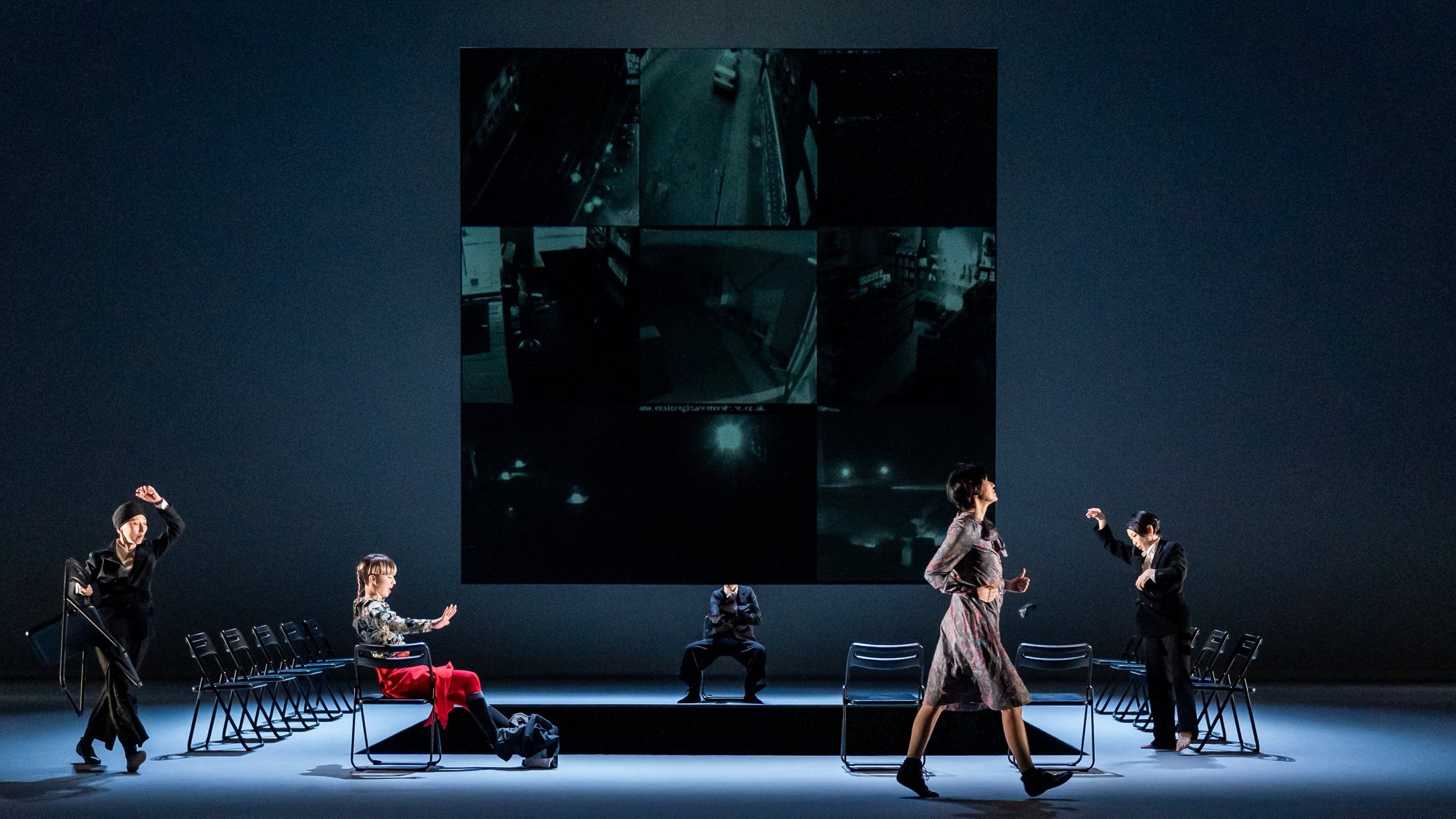 five dancers on stage, a row of black folding chairs, and a large screen displaying a grid of dark surveillance video feeds