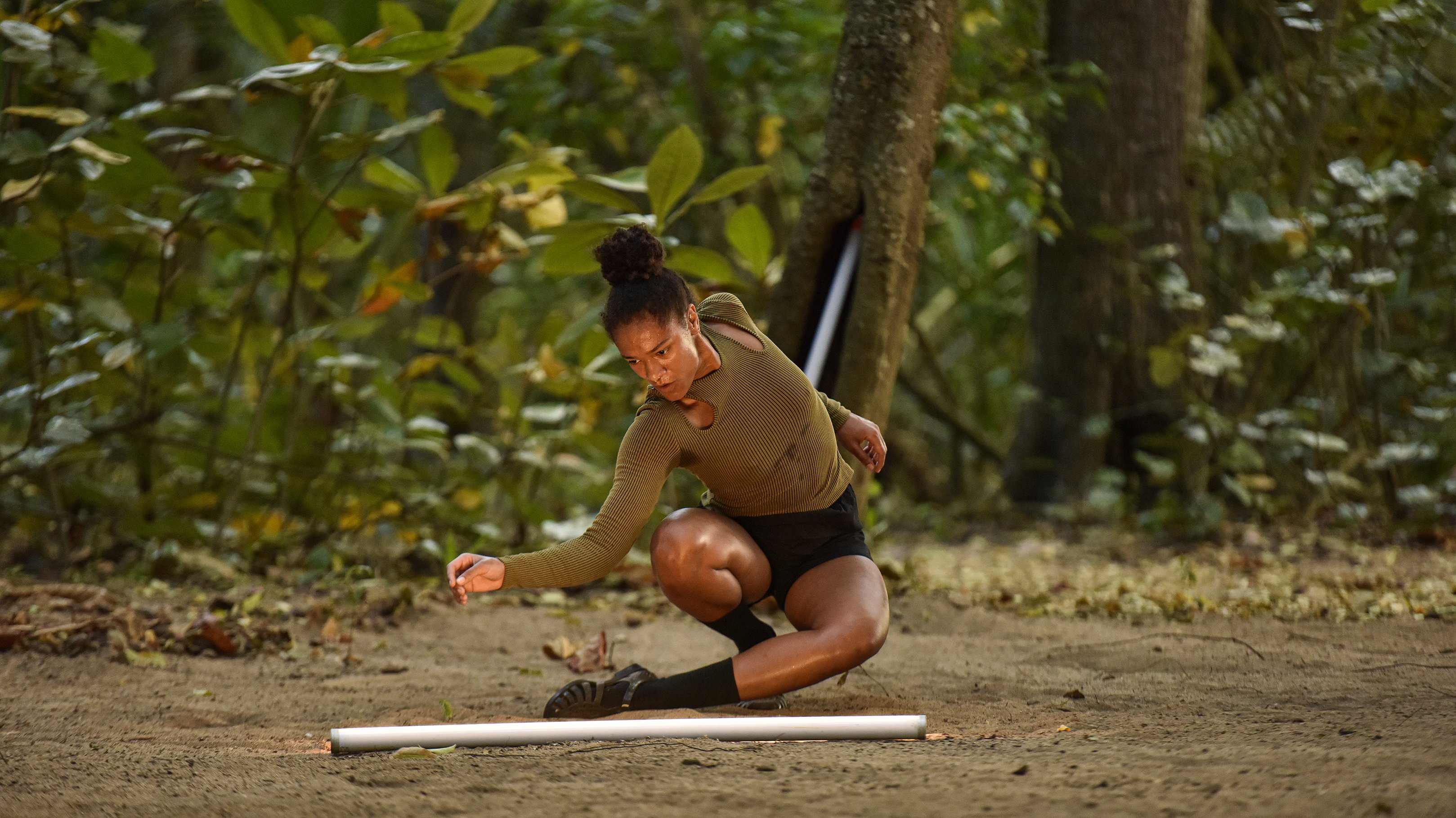 Dancer crouching on earthy ground in a lush forest, arm extended towards a white tube placed on the ground