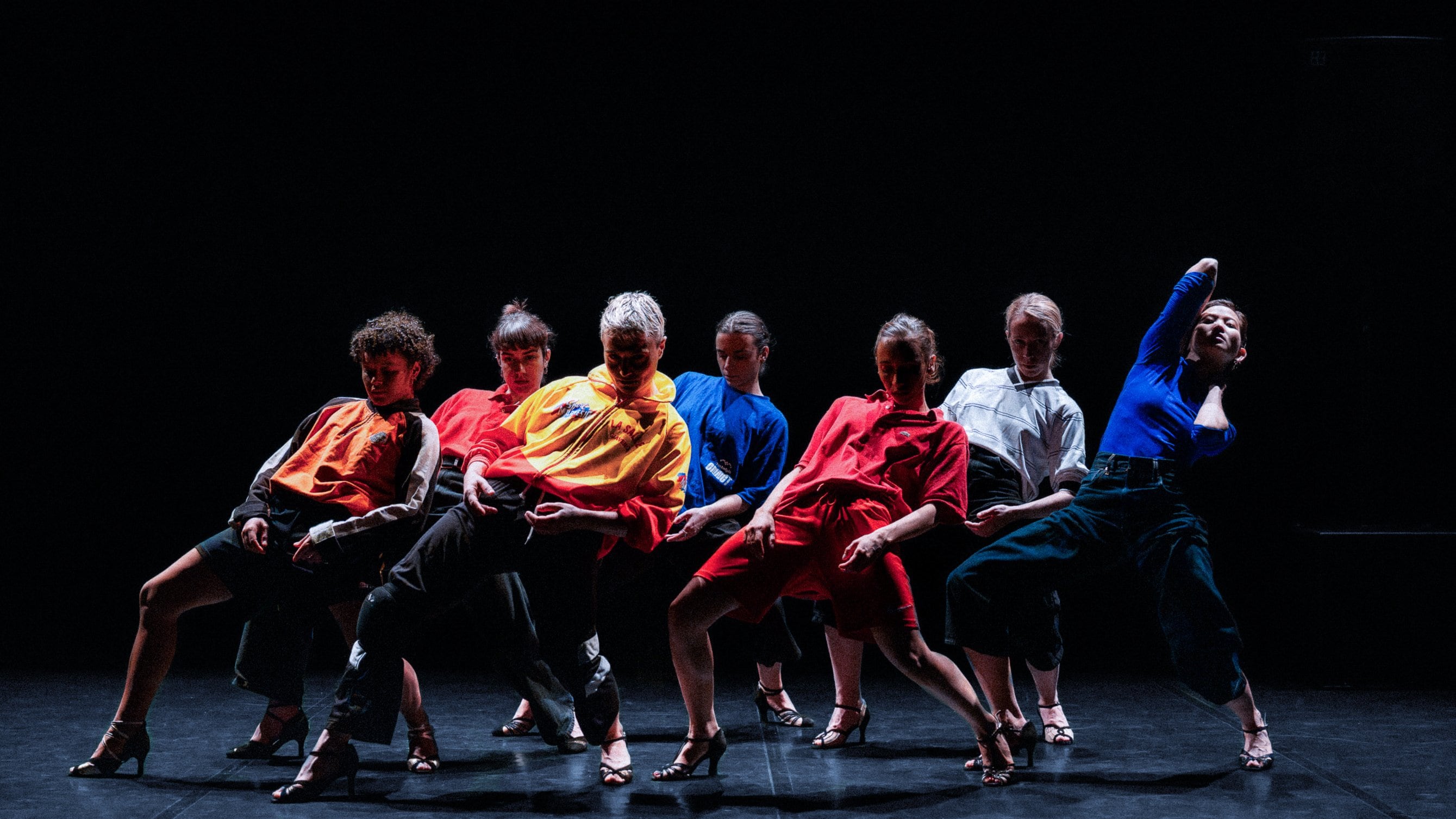 Group of dancers in colorful clothing and high heels striking dynamic poses on a dark stage