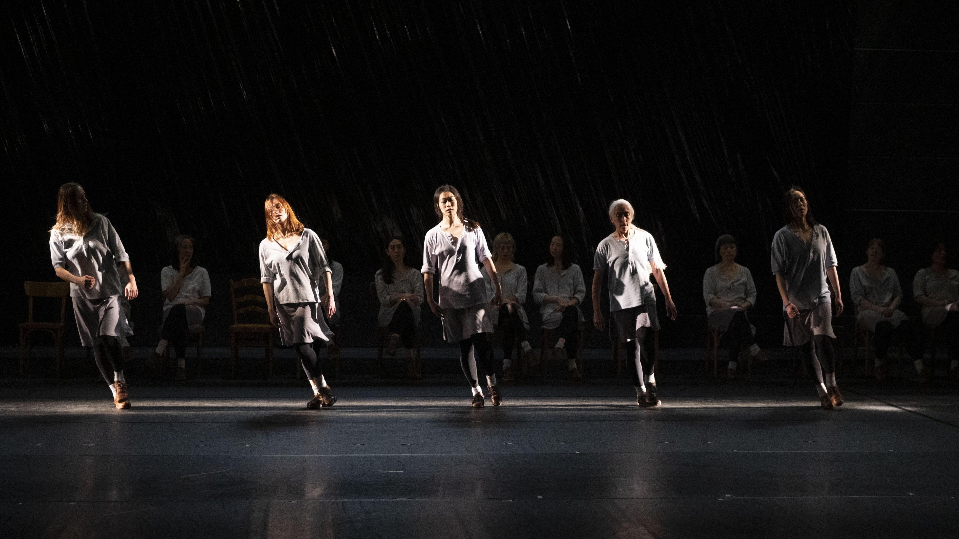 Dancers on a dark stage, some standing in the light, others seated in the shadow, wearing light tunics