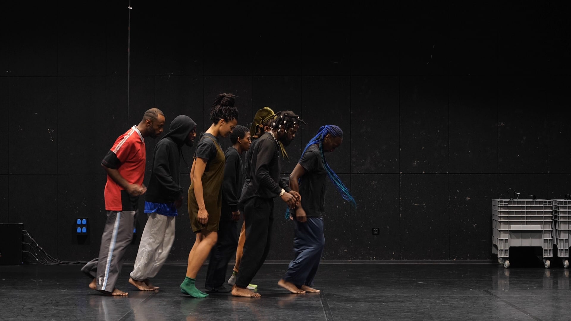 Six dancers, mostly barefoot, moving in a line across a dark stage