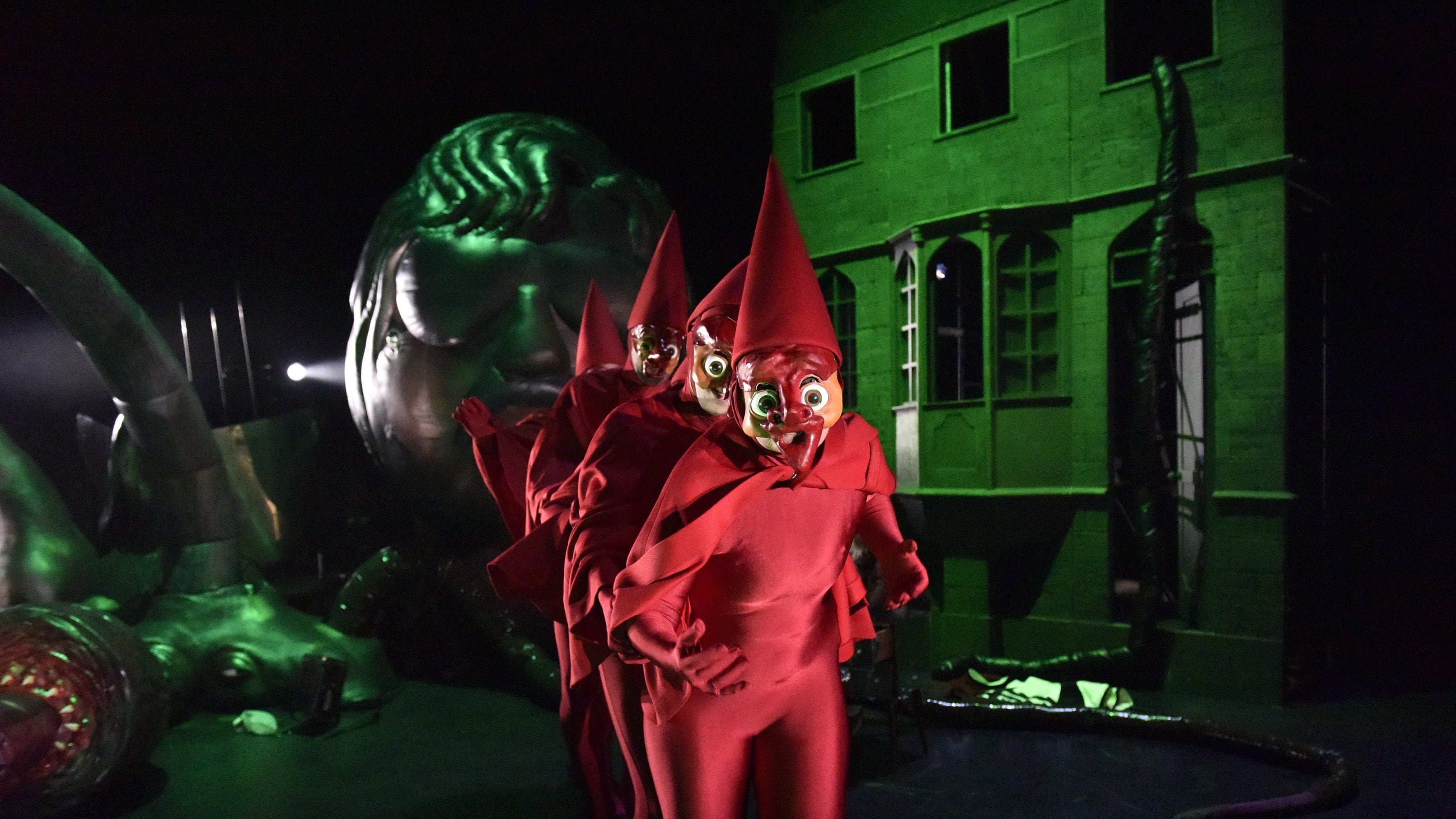 Personnages masqués en costumes rouges vifs et chapeaux pointus, marchant en ligne devant un bâtiment éclairé en vert