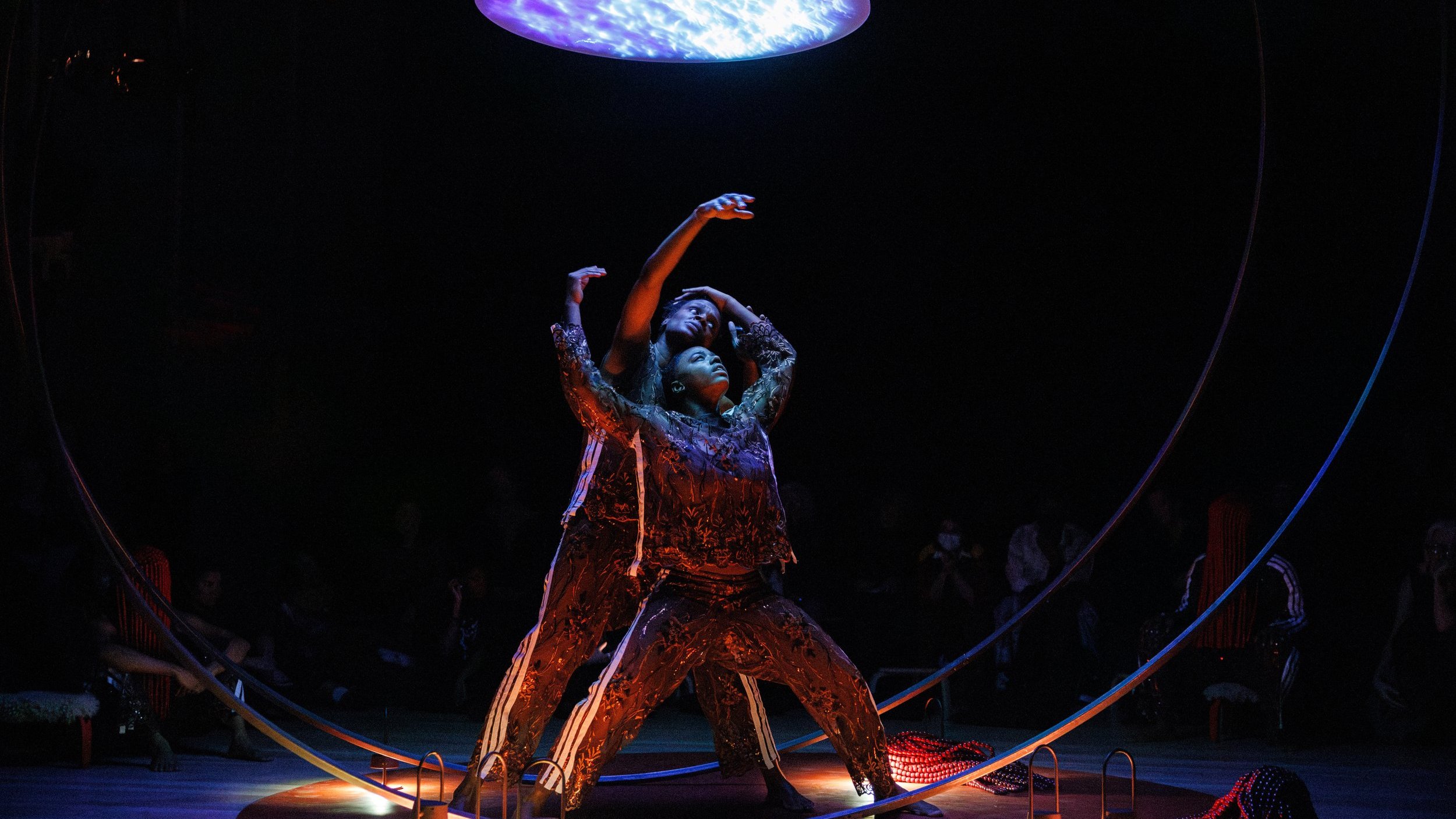 Deux danseurs regardent vers un plafond lumineux, leur corps embrasse un même mouvement dans une sphère métallique. 
